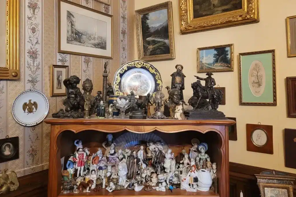 Collection d'objets anciens et de bibelots dans une pièce décorée, incluant sculptures, tableaux, assiettes et figurines en porcelaine.